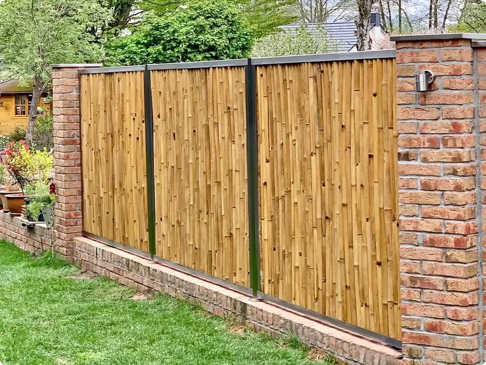 Bambuszaun mit dunklen Metallprofilen zwischen Ziegelpfeilern als Sichtschutz in einem bepflanzten Garten.