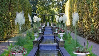 Stufenförmiger Springbrunnen in einem gepflegten Garten, umgeben von hohen Grasbüscheln und Hecken. Entlang des Brunnens stehen weiße Pflanzkübel mit roten Blumen. Im Hintergrund Pergola und Bäume, die eine ruhige, harmonische Atmosphäre schaffen.