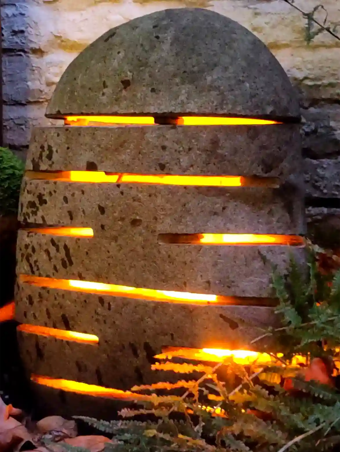 Steinlampe aus indonesischem Flussstein Steinlampe mit waagerechten Schlitzen und warmen Lichtschein im Garten bei Nacht.