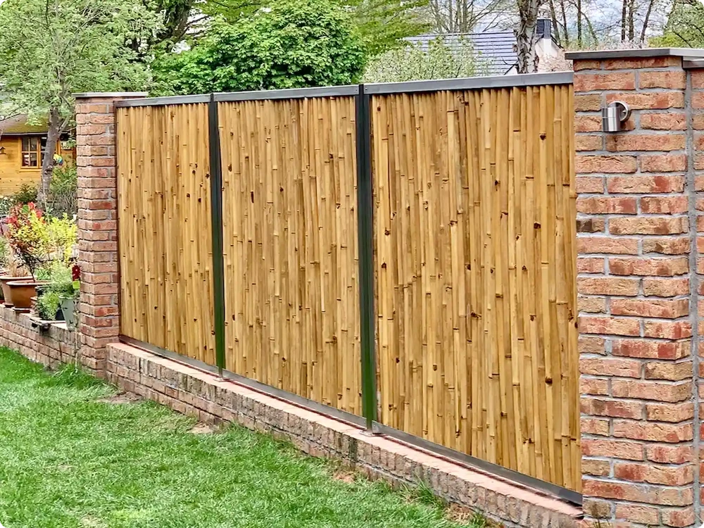 Bambuszaun mit dunklen Metallprofilen zwischen Ziegelpfeilern als Sichtschutz in einem bepflanzten Garten.