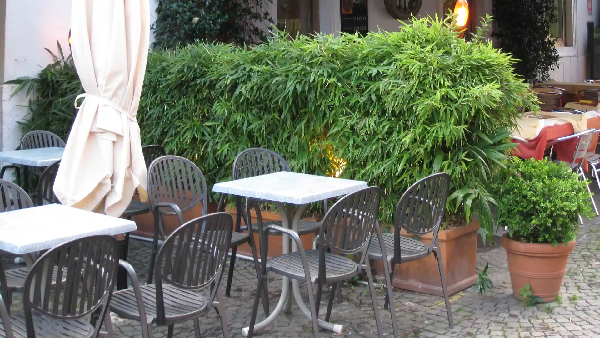 Freiluftcafé mit Metalltischen und -stühlen, geschlossener Sonnenschirm; umgeben von üppigen grünen Hecken in Pflanzgefäßen auf Kopfsteinpflaster; im Hintergrund weitere Tische mit Tischdecke.