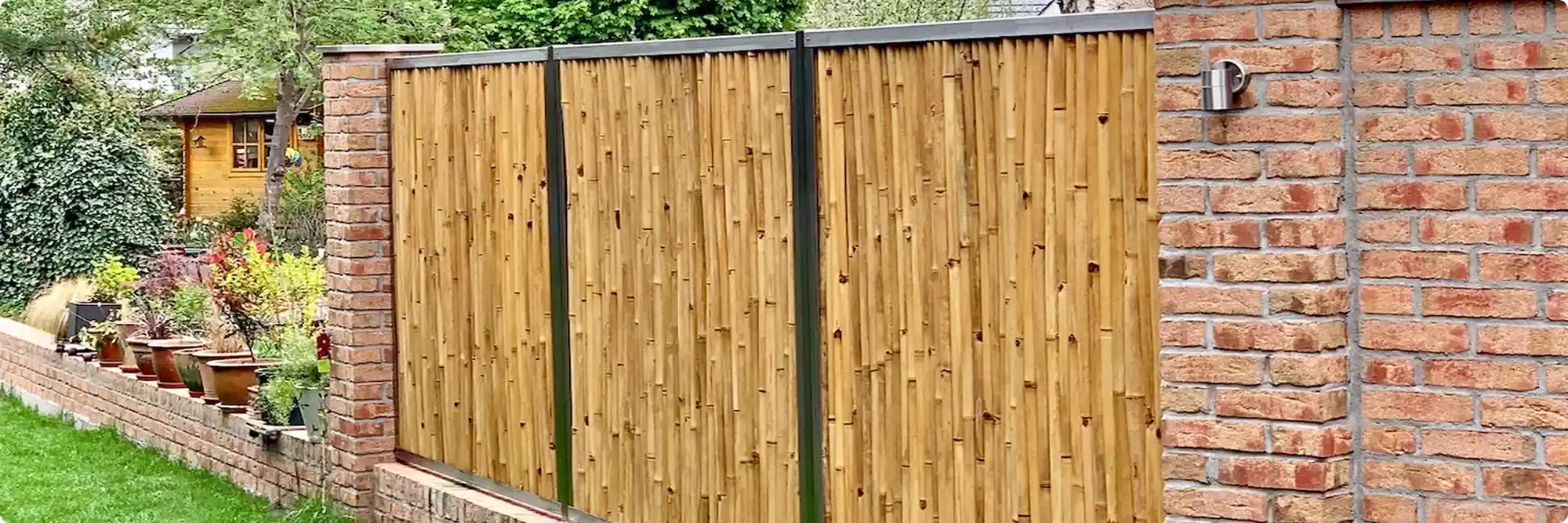 Bambuszaun mit dunklen Metallprofilen zwischen Ziegelpfeilern als Sichtschutz in einem bepflanzten Garten.